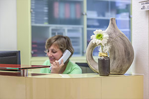 Deko-Vase mit Blume im Vordergrund auf dem Empfangstresen, im Hintergrund telefoniert eine Praxis-Mitarbeiterin.
