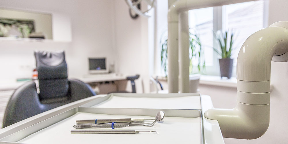 Sterile zahnärztliche Instrumente auf einem Tablett im Vordergrund, im Hintergrund ist ein Behandlungsstuhl und Fenster zu sehen.
