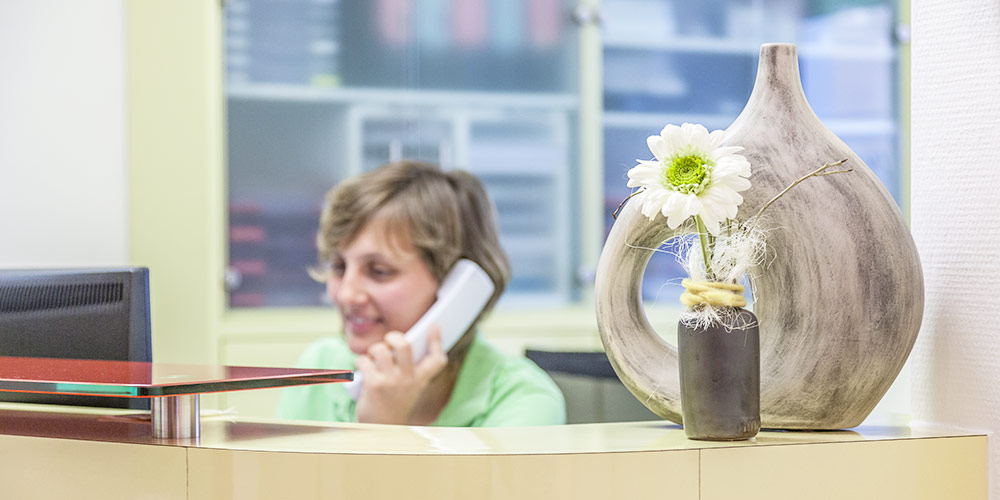 Rezeption einer Arztpraxis mit dekorativer Vase und einer einzelnen Blume im Vordergrund; im unscharfen Hintergrund telefoniert eine Mitarbeiterin.